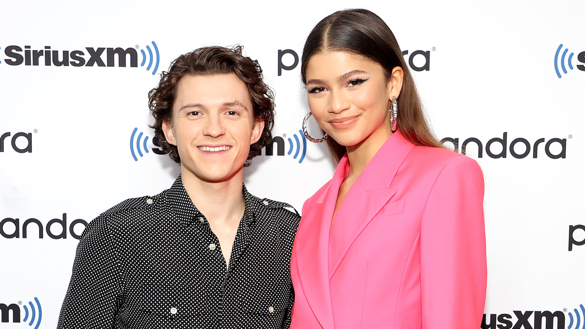 Tom Holland and Zendaya pose together at a SiriusXM event. Tom is in a black shirt, and Zendaya wears a bright pink suit.