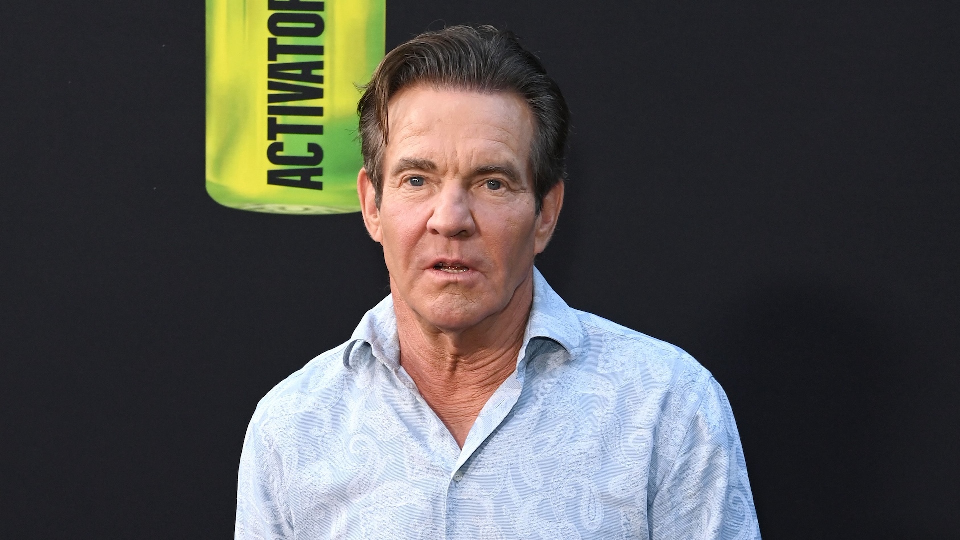 Dennis Quaid wearing a paisley button-up shirt standing against a black backdrop.