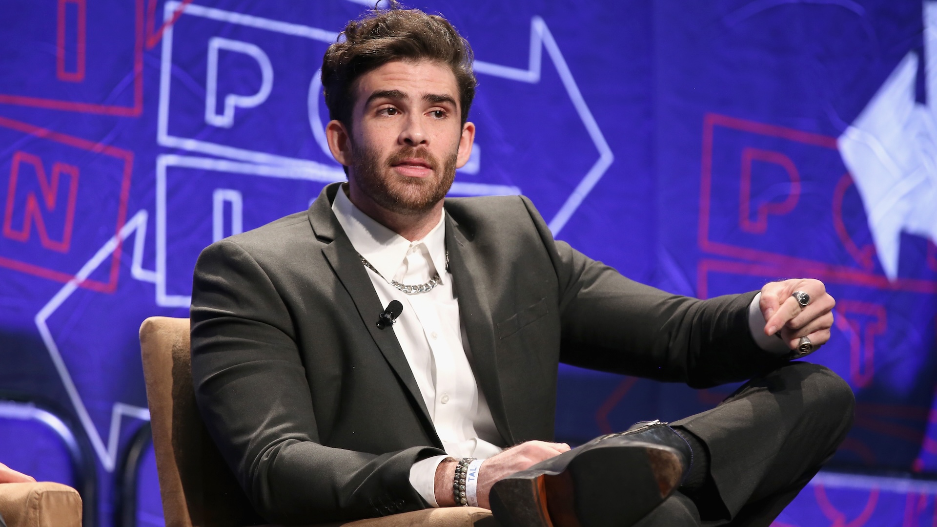 Hasan Piker wearing a black suit sitting against a blue backdrop.