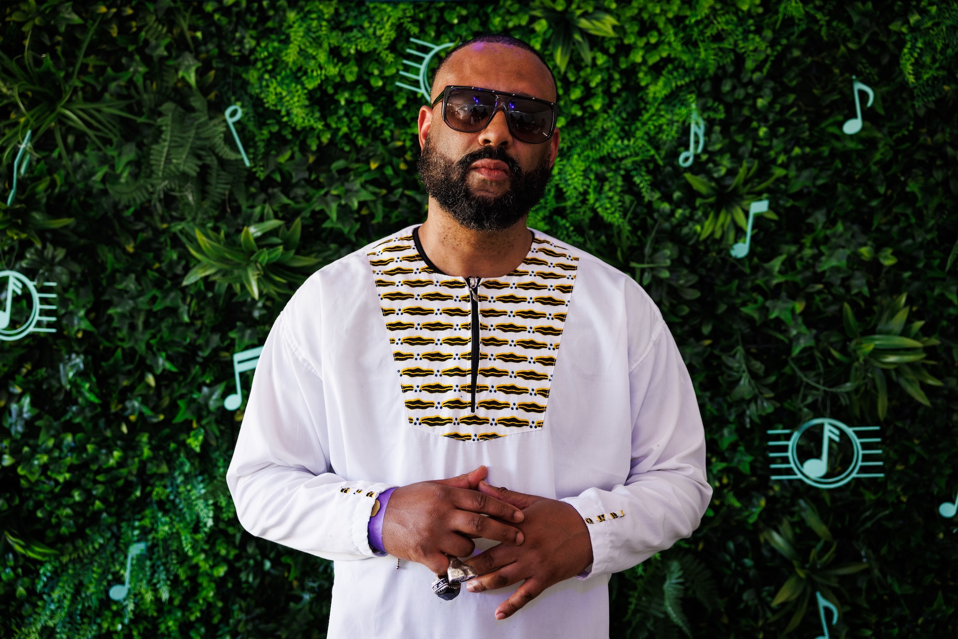 Madlib with a beard and sunglasses stands in front of a green leafy background, wearing a white shirt with a patterned design.