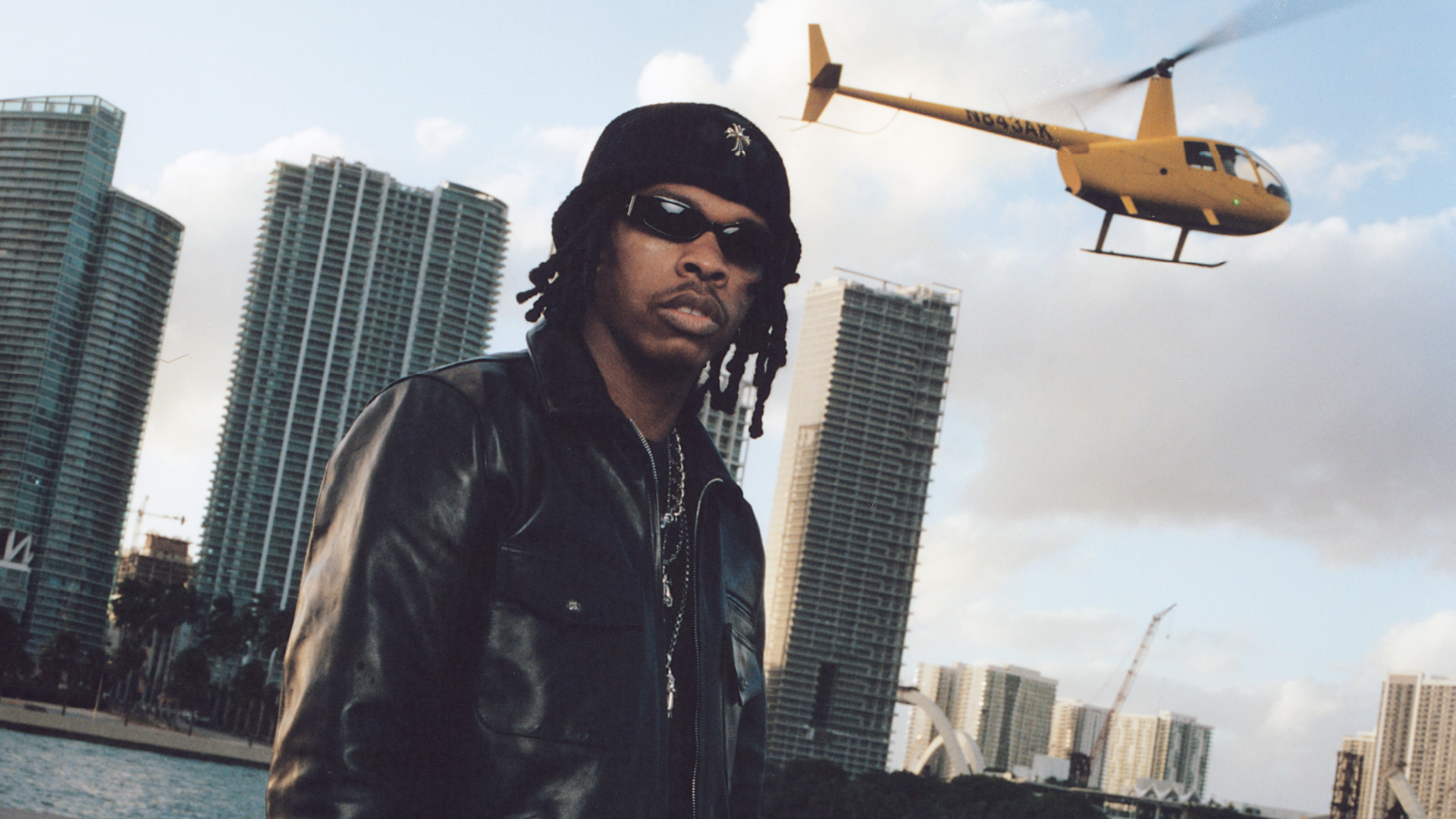 A man in a black leather jacket and sunglasses stands in front of city skyscrapers. A yellow helicopter flies overhead.