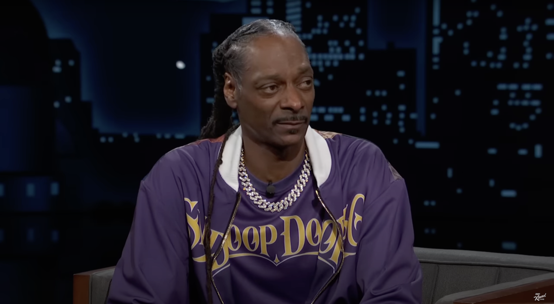 Snoop Dogg wearing a purple "Snoop Dogg" shirt and chain necklace, sitting on a talk show set.