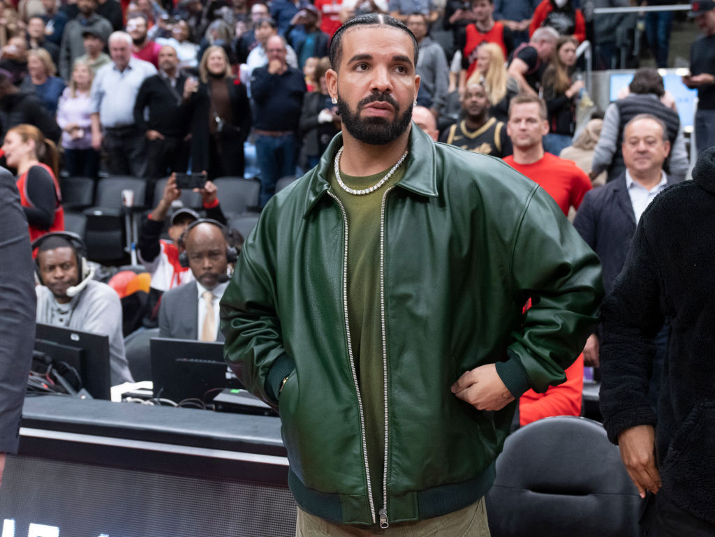 Drake in a green jacket at a basketball game, surrounded by fans and spectators in the background.