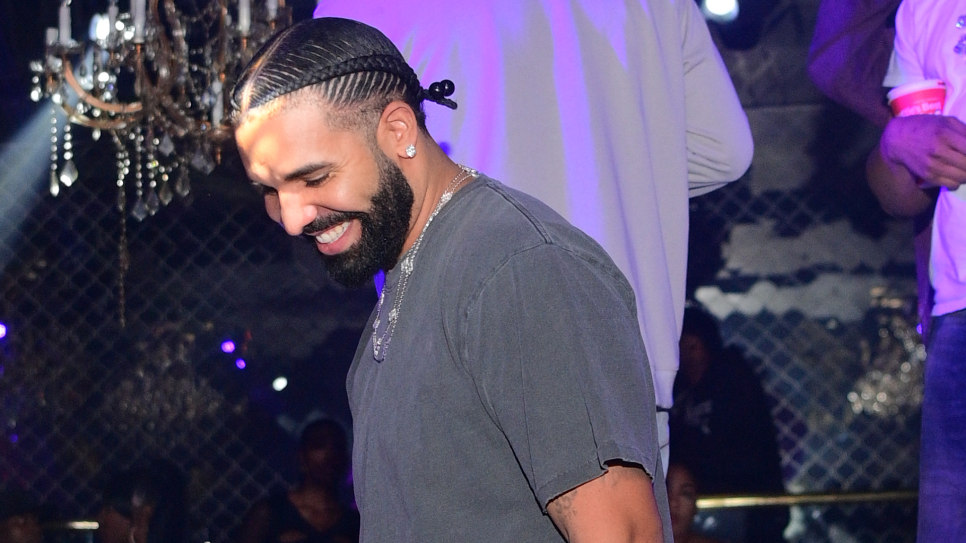 Drake smiling in a nightclub, wearing a dark shirt with braided hair. Chandelier and people in the background.