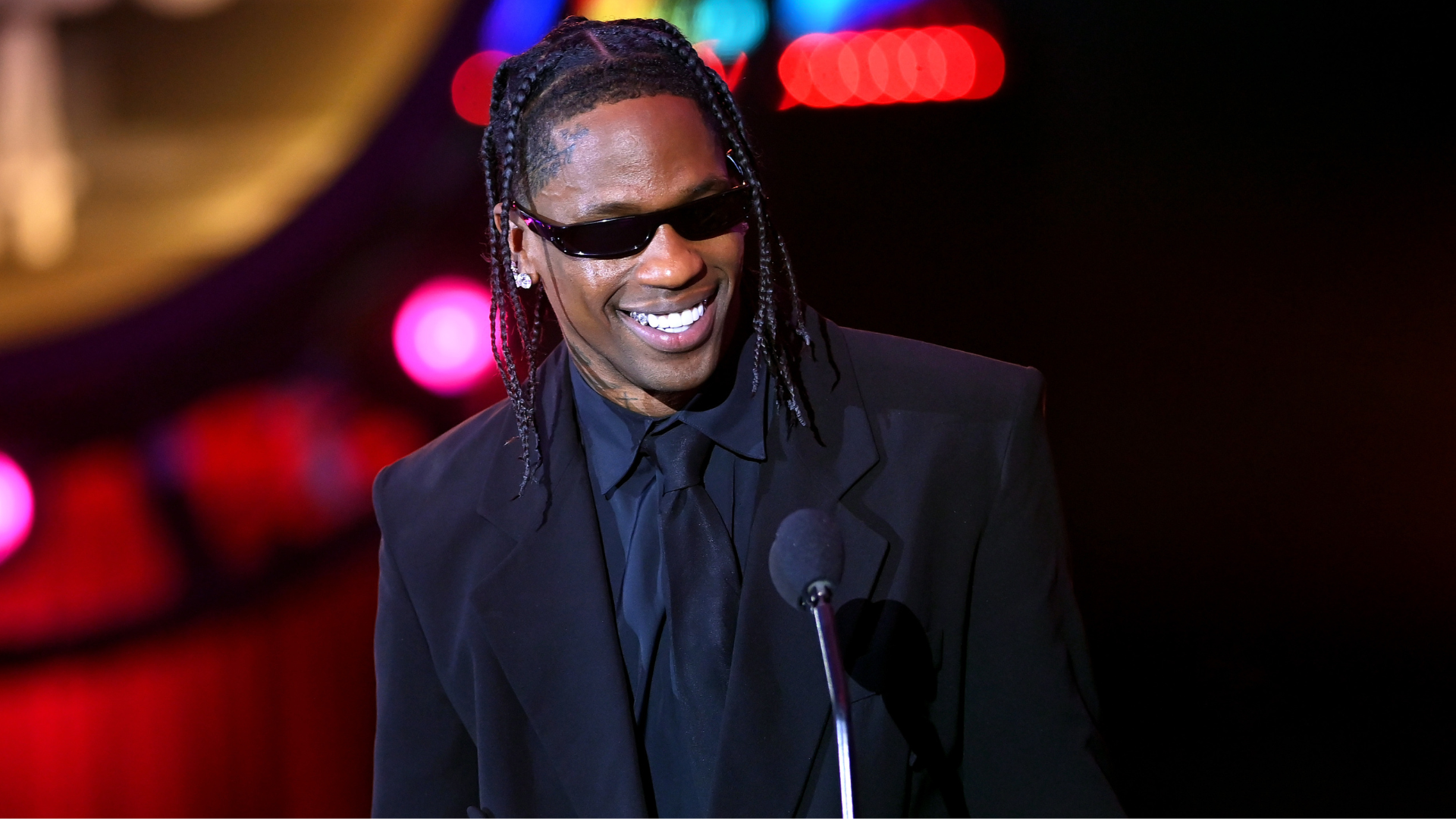 Travis Scott in sunglasses and a black suit, smiling at a microphone on stage.