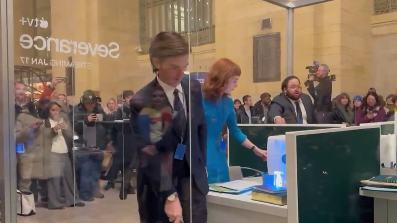 A scene from an event for the Apple TV+ show "Severance," featuring actors in an office setup with an audience watching through glass.