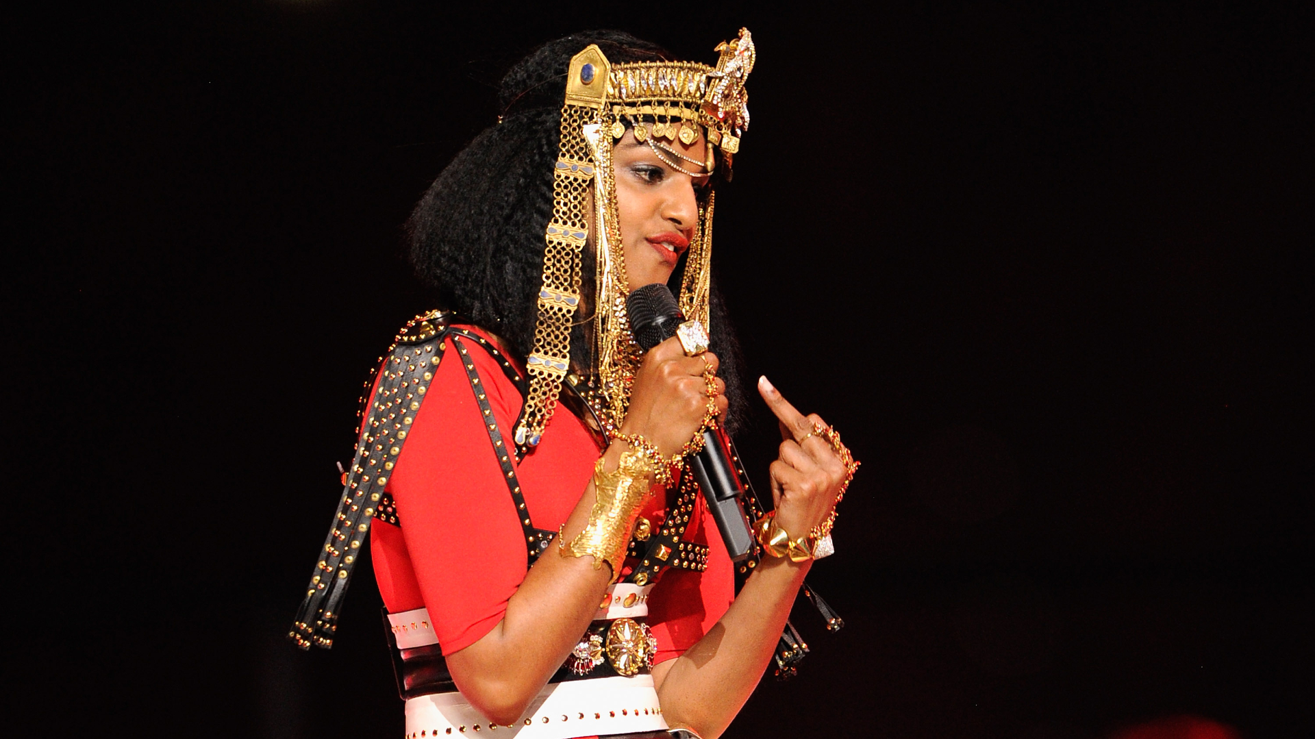 M.I.A. performing, dressed in an Egyptian-inspired outfit with gold accessories, holding a microphone.