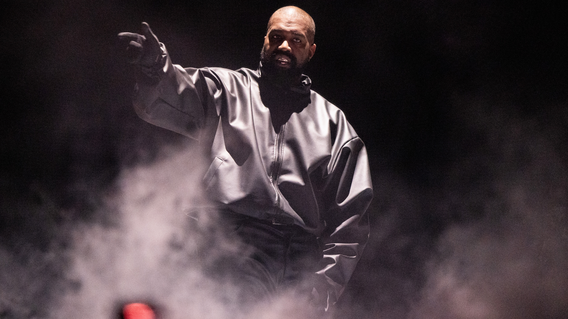 Ye performing on stage, wearing a black jacket amidst smoke and dramatic lighting.