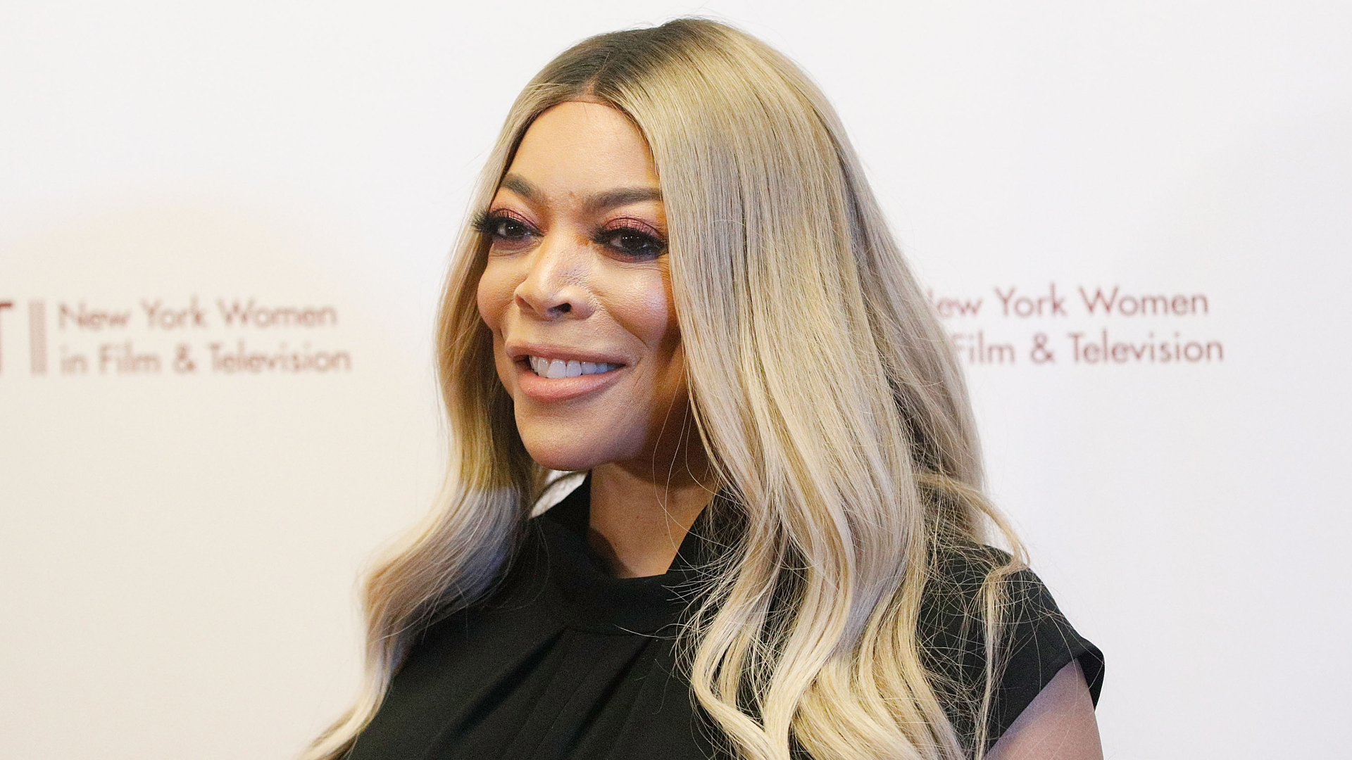 Wendy Williams smiling at an event, wearing a black top, with long blonde hair. Background shows a New York Women in Film & Television logo.