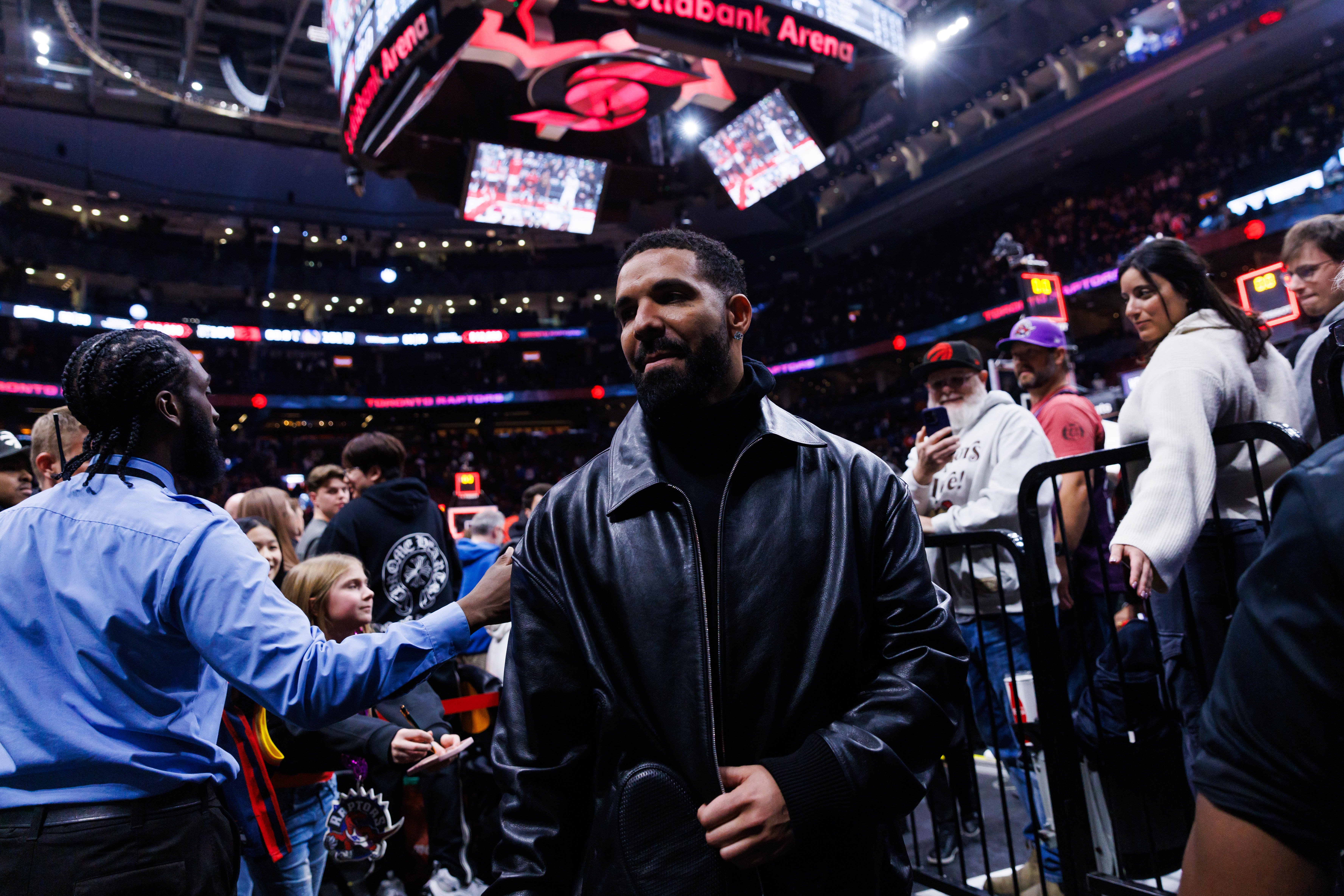Drake in a black leather jacket at Scotiabank Arena, surrounded by fans and security during a basketball game.