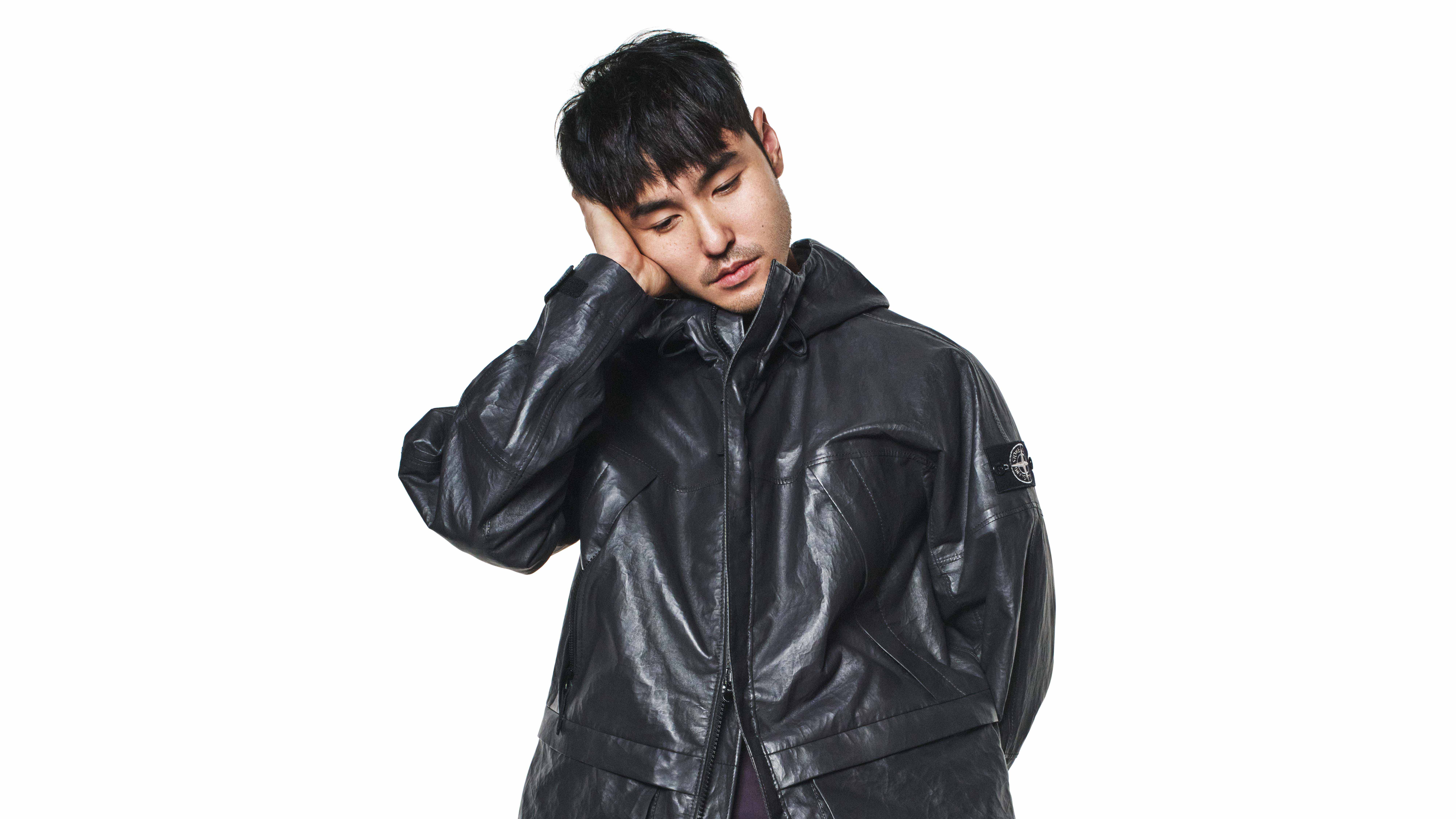 A person in a black leather jacket with a hood, resting their head on their hand, against a plain white background.