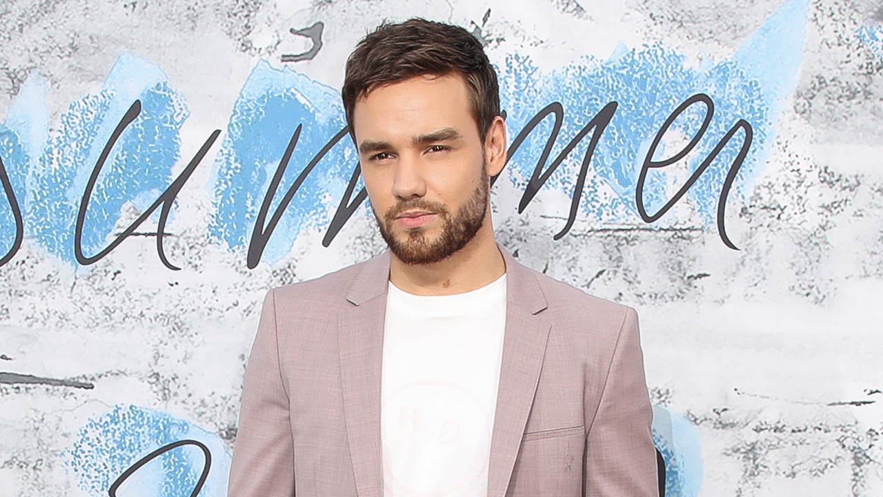 Liam Payne in a light pink blazer and white shirt, standing against a blue and white background.