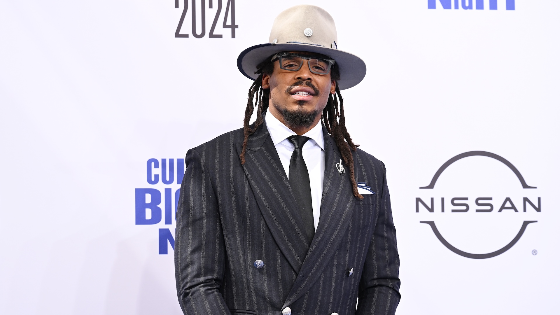 Cam Newton in a pinstripe suit and wide-brimmed hat stands in front of a backdrop with logos, including Nissan, at an event.