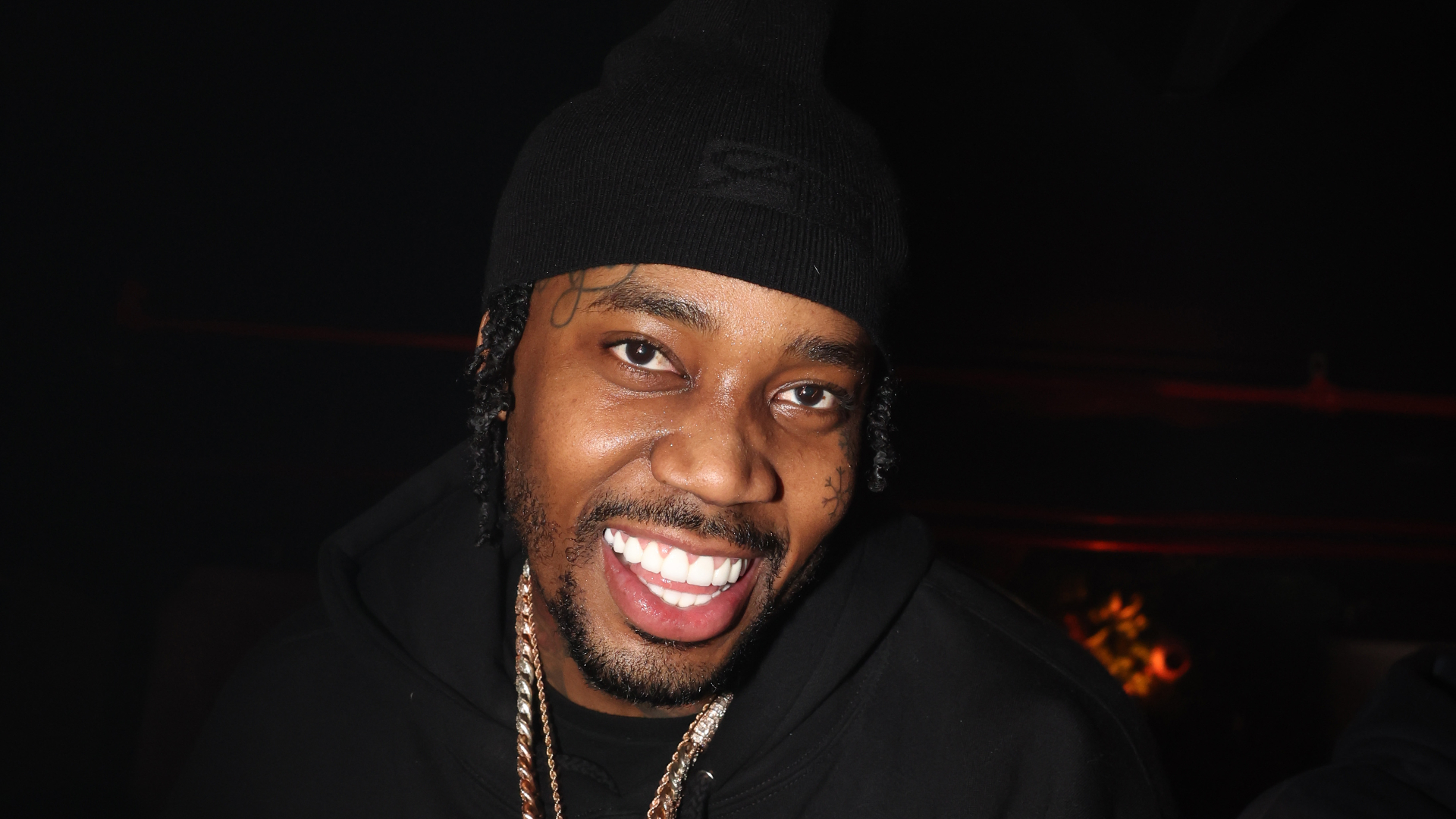 Fivio Foreign wearing a black beanie and hoodie, smiling brightly, with a gold chain visible around their neck.