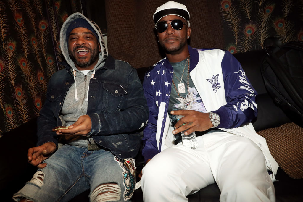 Two men are sitting on a couch. One wears a denim jacket and beanie, the other a white cap and sunglasses. They're both smiling.