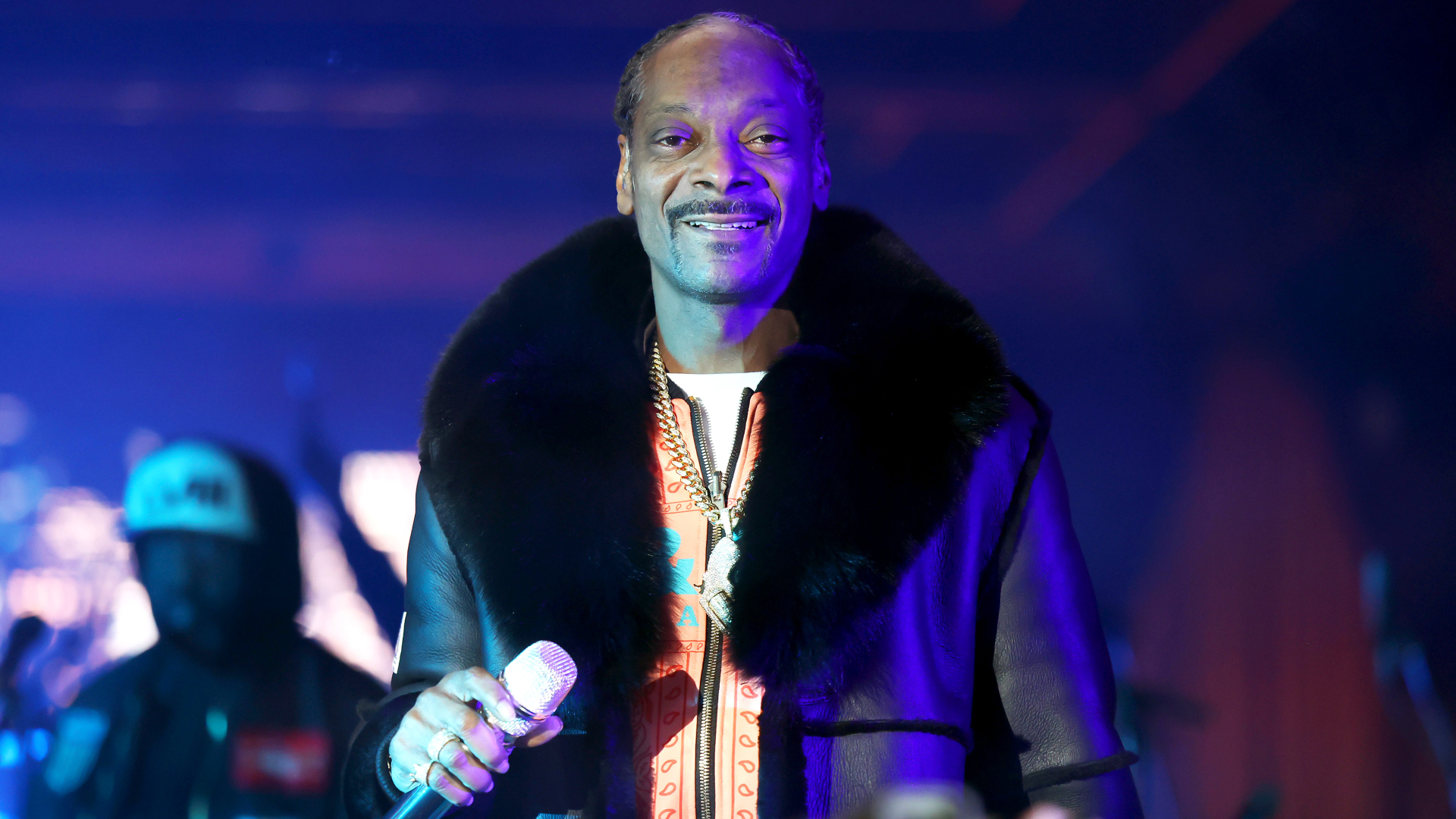 Snoop Dogg performing on stage, wearing a fur-collared coat, holding a microphone, smiling under stage lights.