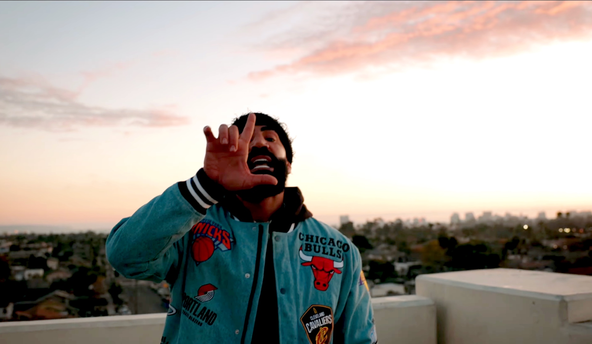 Streamer Yousef “Fousey” Erakat, now going by "Crashout YSE," stands on top of a building, making an L-shape with his right hand while wearing a turquoise jacket with various basketball team logos on it.