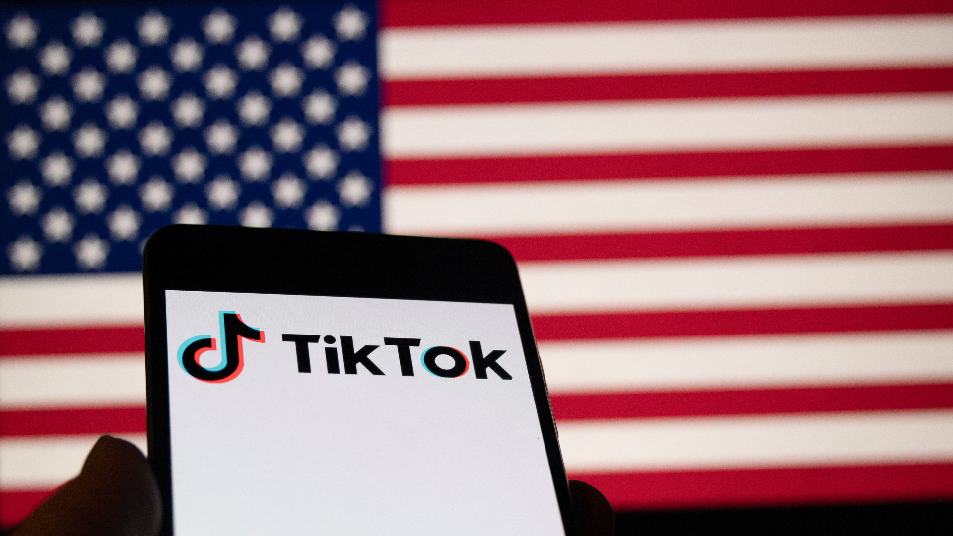 A smartphone displaying the TikTok logo in front of a blurred American flag background.