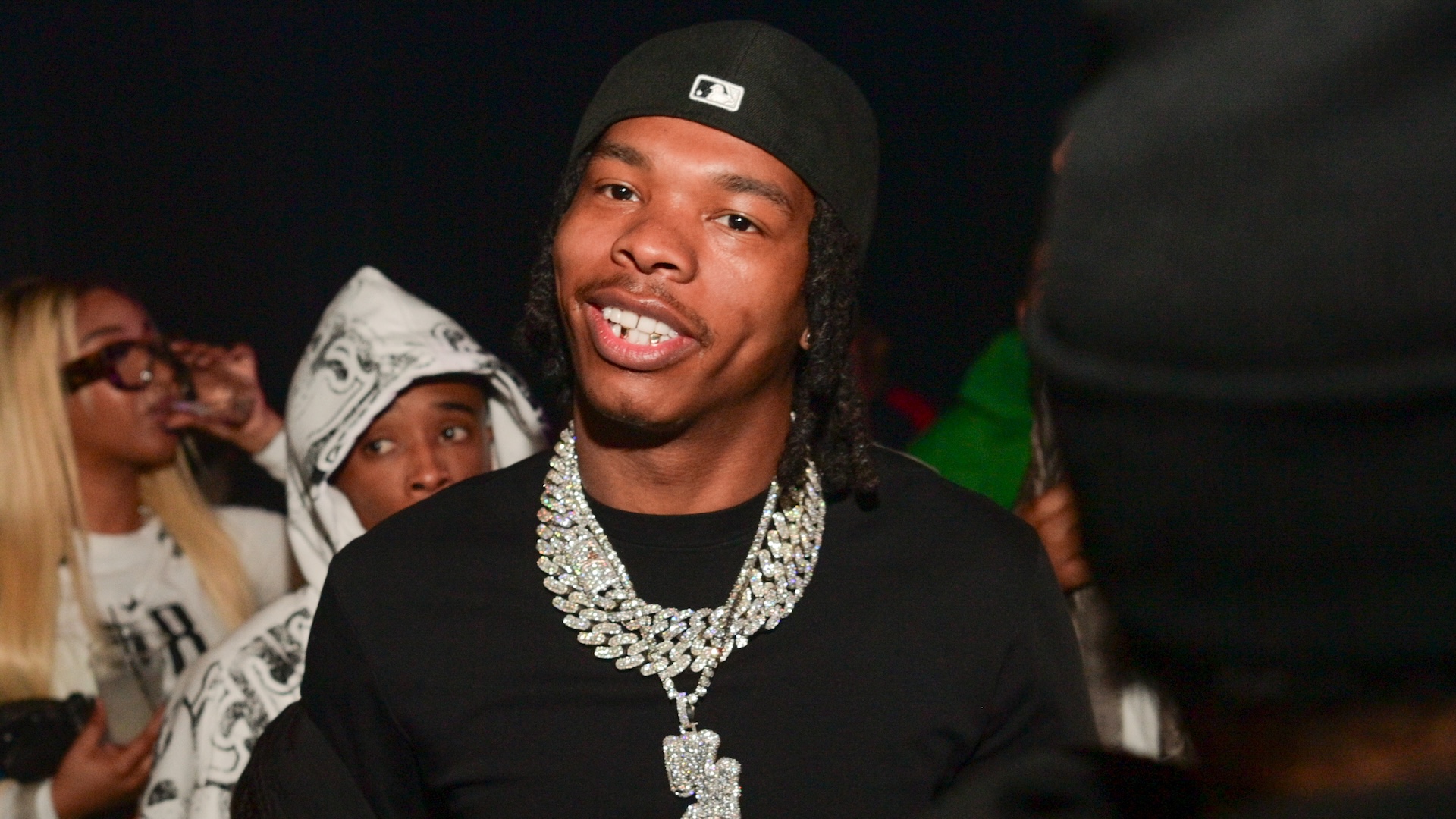 Lil Baby, wearing a black cap and diamond necklace, smiles at an event with people in the background.