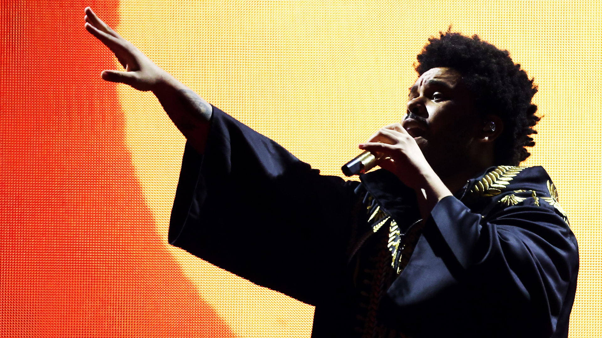 The Weeknd performing on stage, wearing a dark robe with gold details, holding a microphone, against a warm, orange-lit background.