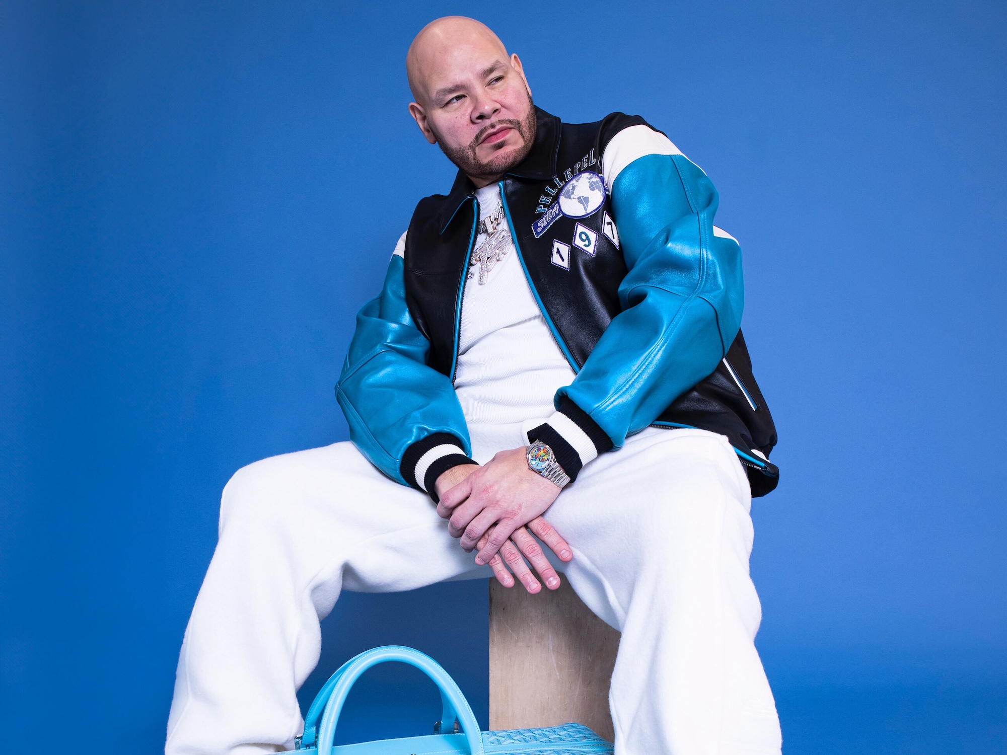 Fat Joe sitting against a blue background, wearing a black and teal jacket, white pants, and a watch, with a blue bag nearby.