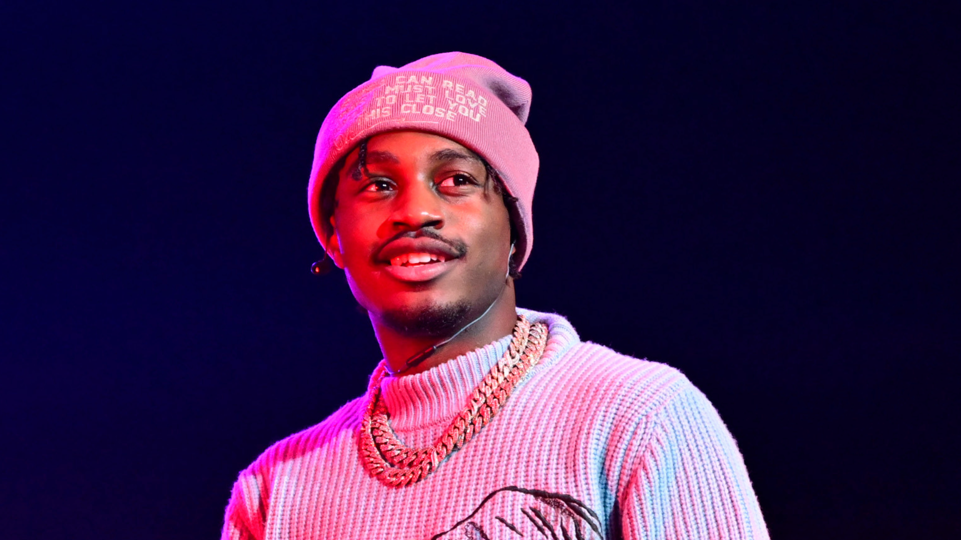 A man wearing a pink beanie and a sweater, with a gold chain, stands against a dark background, illuminated by pink and blue lights.