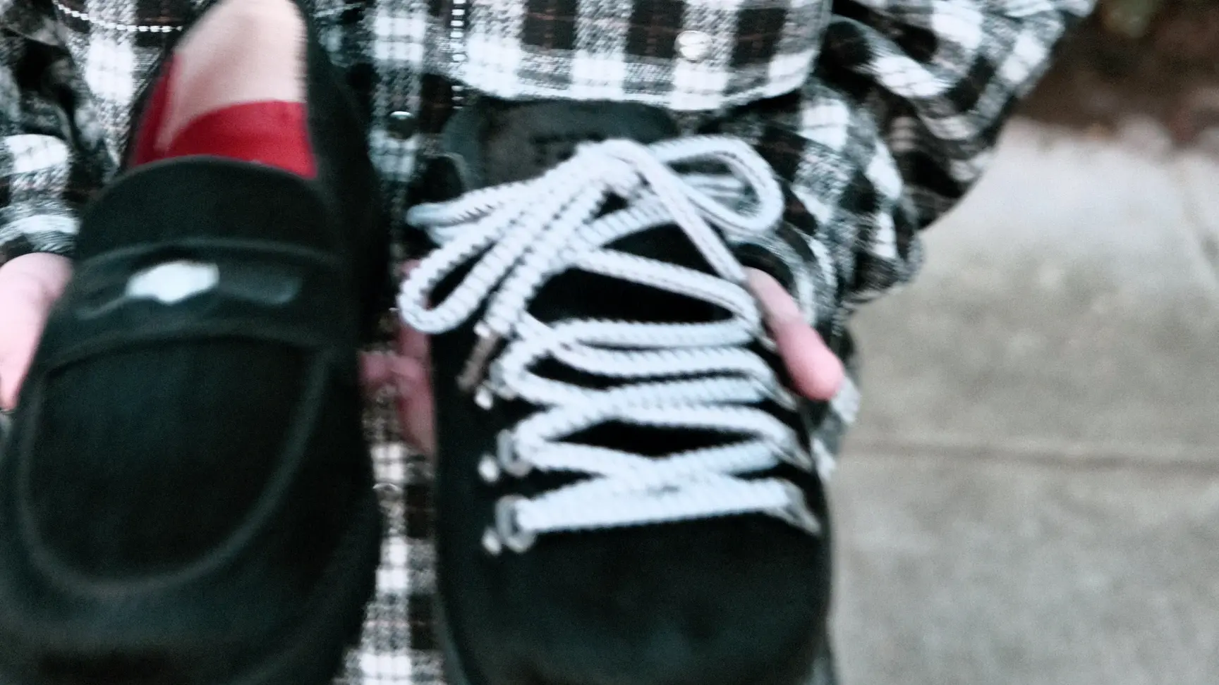 A person in a plaid shirt holds two black shoes, one with laces and one without, on a sidewalk.