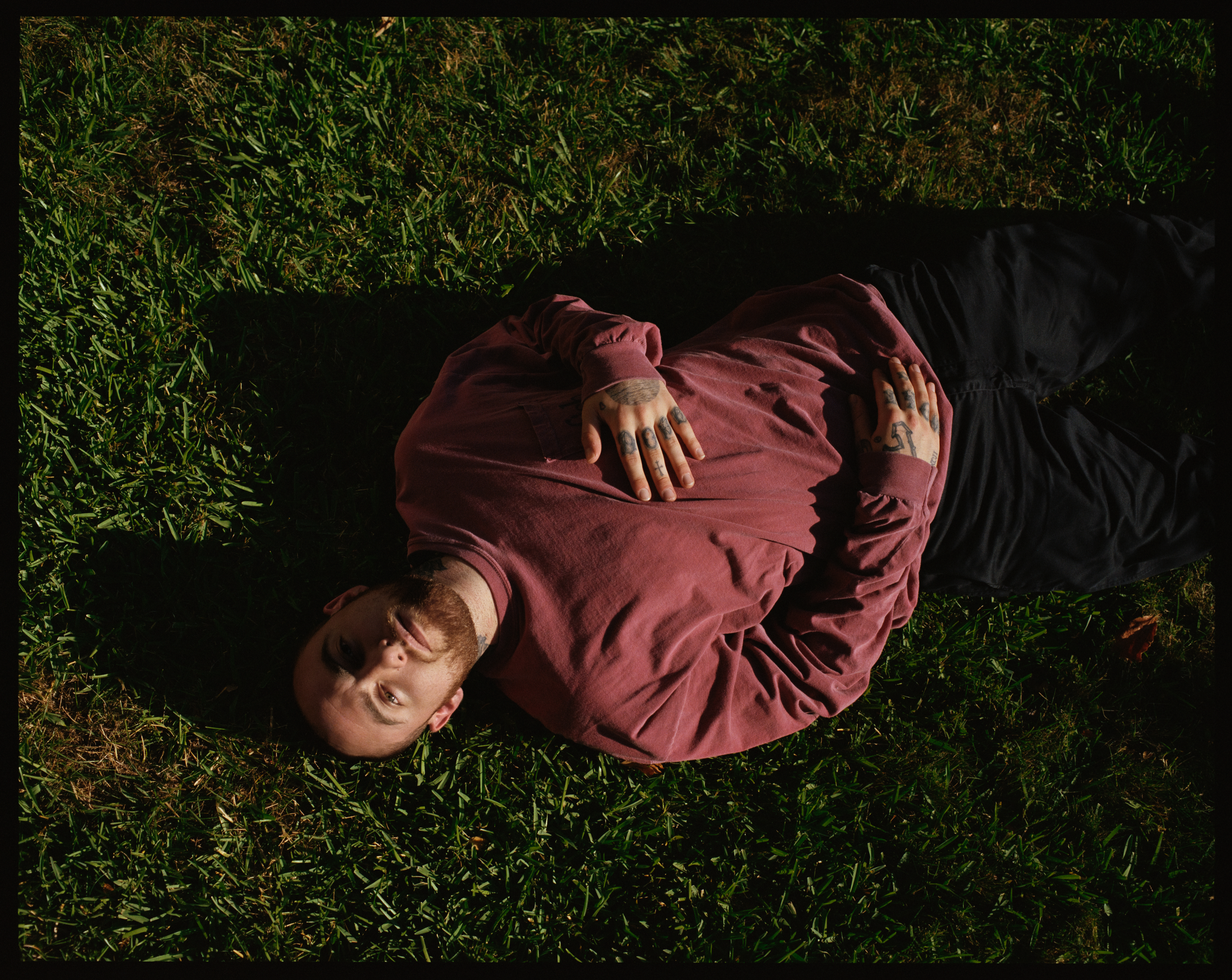 Mac Miller on grass wearing a maroon shirt and black pants, with tattoos on their hands.