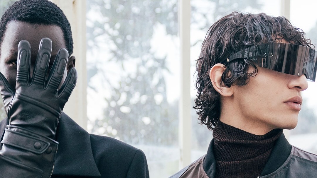 Two models in stylish outfits; one covers their face with a gloved hand, the other wears futuristic sunglasses.