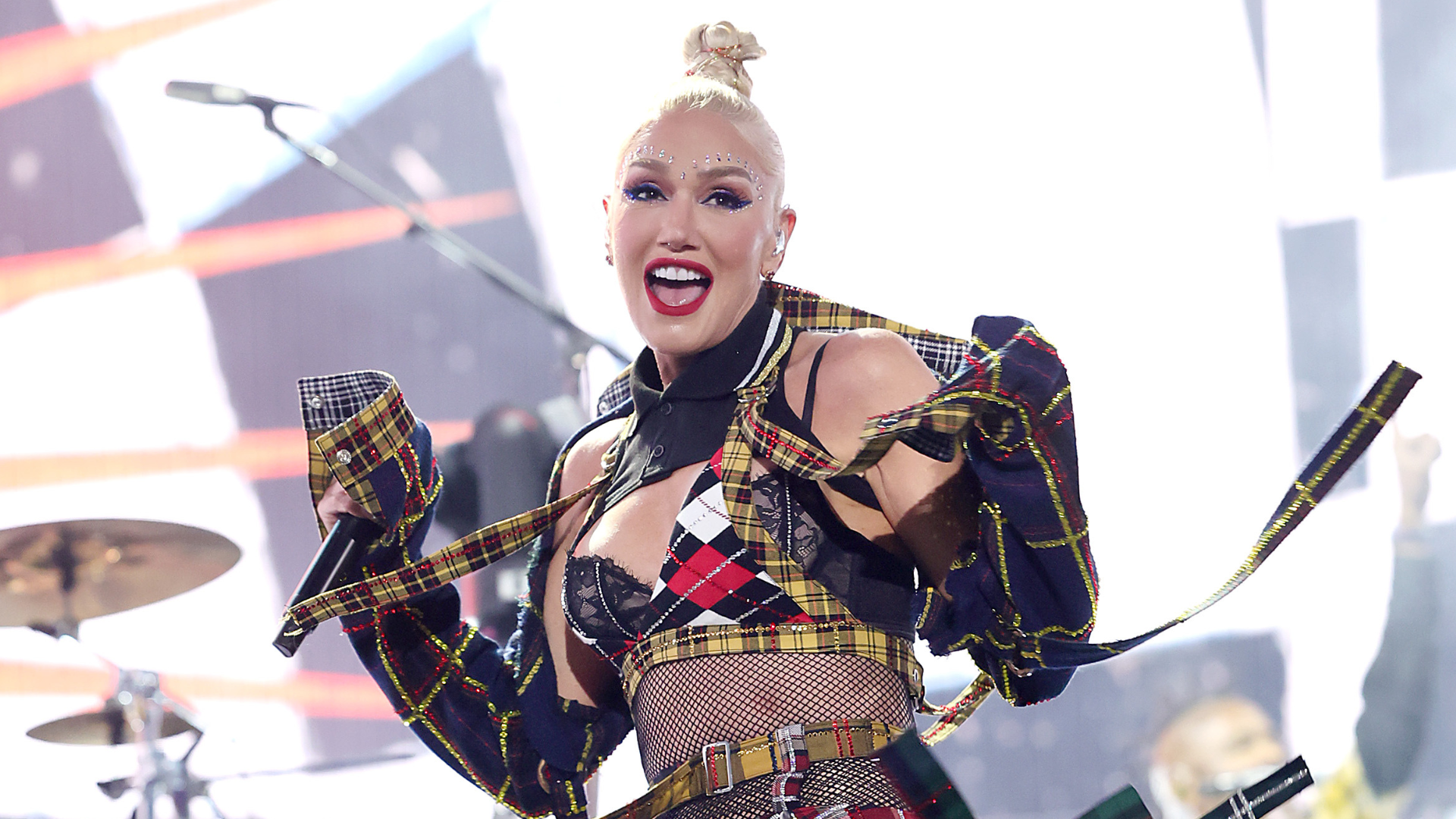 Gwen Stefani performing on stage, wearing a plaid outfit with bold makeup and a high bun, smiling energetically.