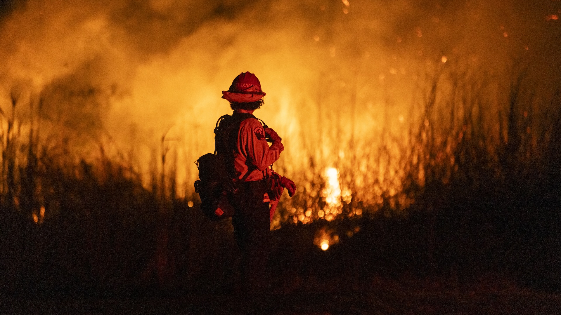 A firefighter in protective gear stands in front of a large blaze, with flames and smoke illuminating the night sky.