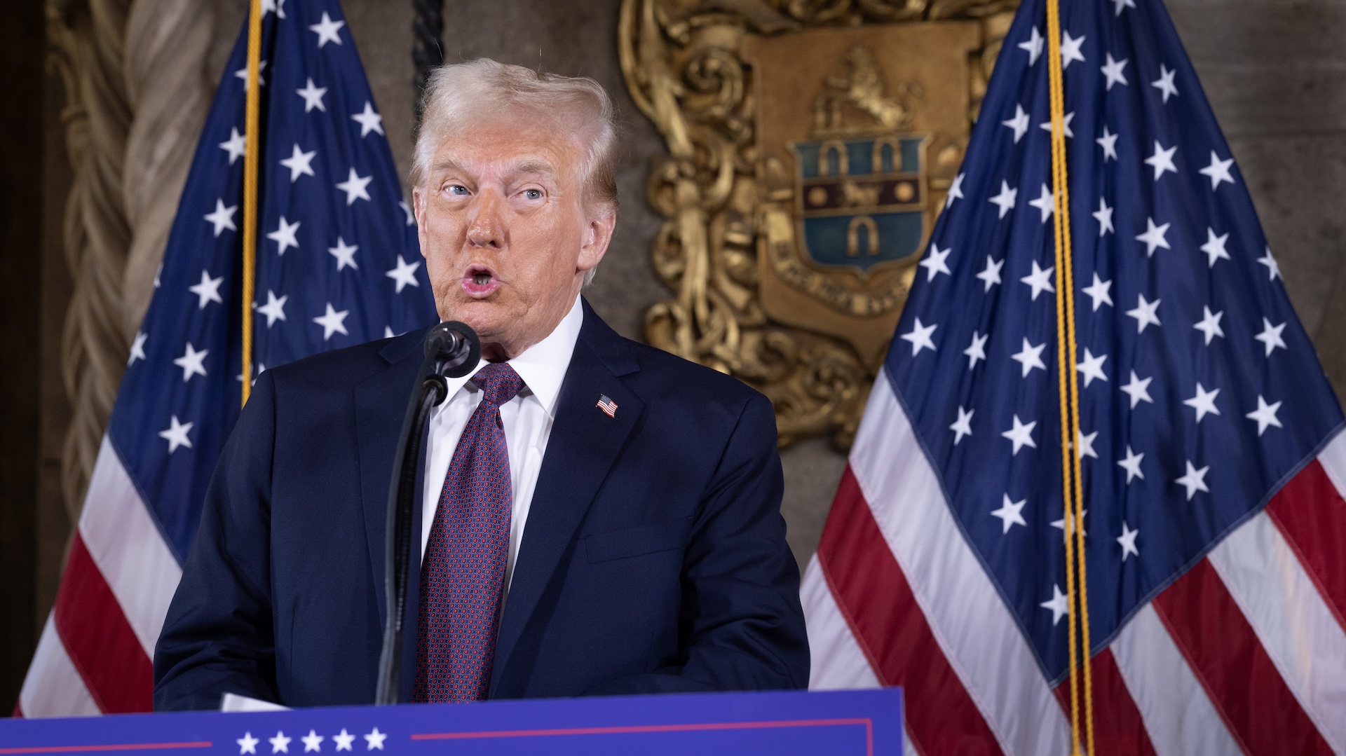 Donald Trump speaking at a podium with two American flags behind him, wearing a dark suit and red tie.