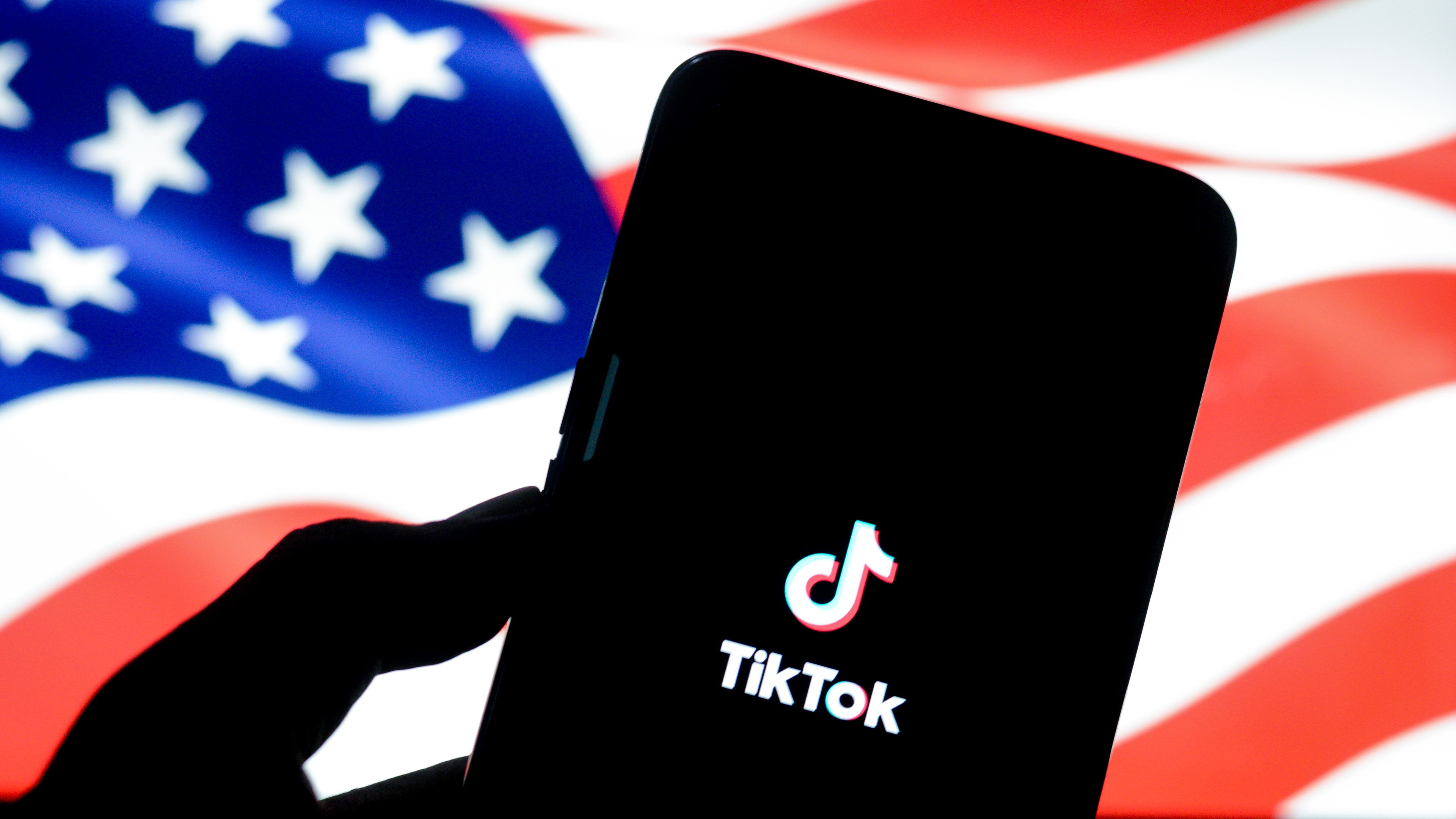 A smartphone with the TikTok logo is held up against a blurred background of an American flag.