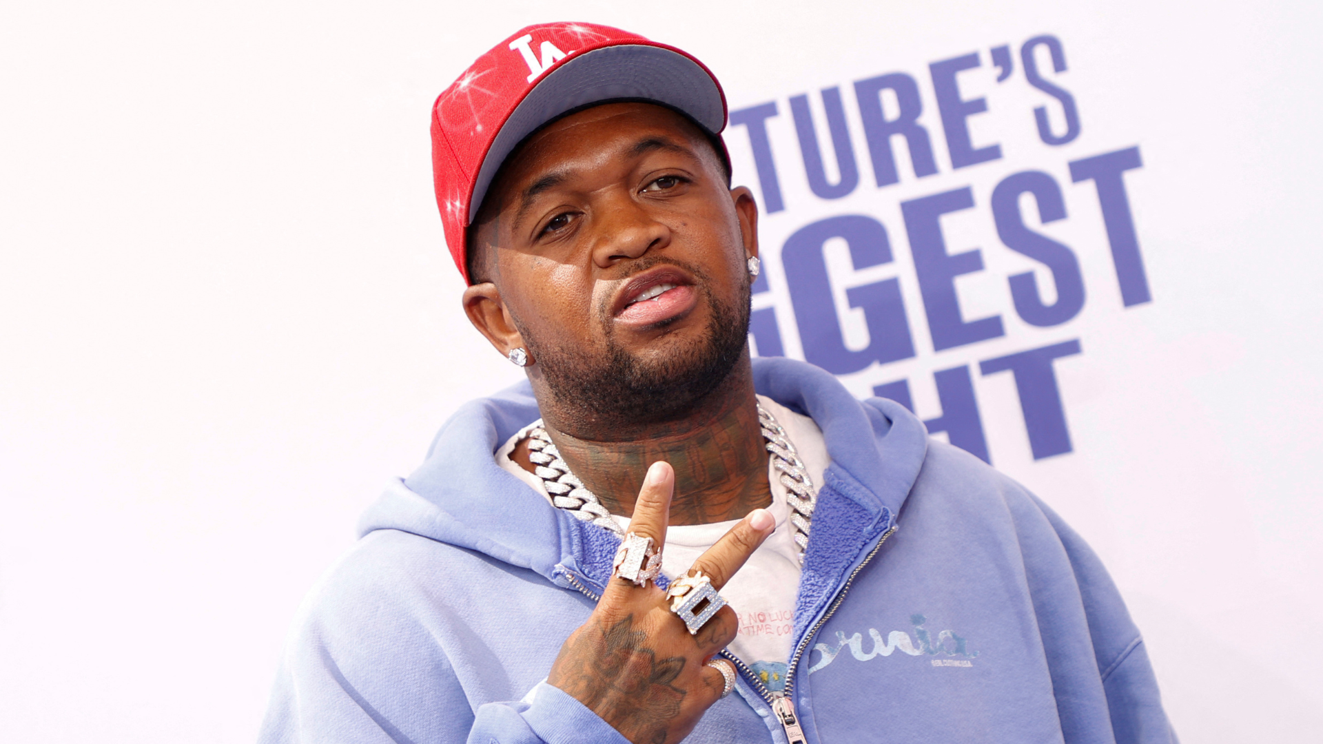 DJ Mustard wearing a red cap and blue hoodie, making a peace sign.