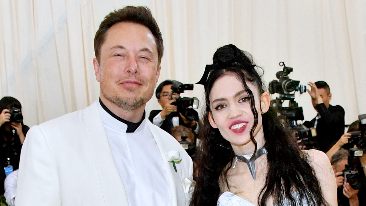 Elon Musk and Grimes at an event, smiling and posing for photos. Elon is in a white suit, and Grimes is wearing a unique dress.