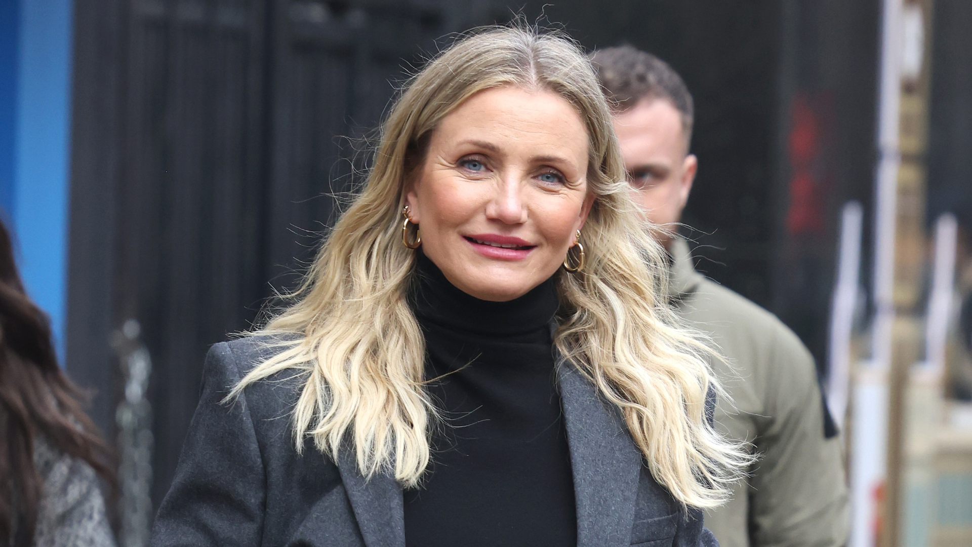 Cameron Diaz with long blonde hair, wearing a black turtleneck and grey coat, smiling outdoors.