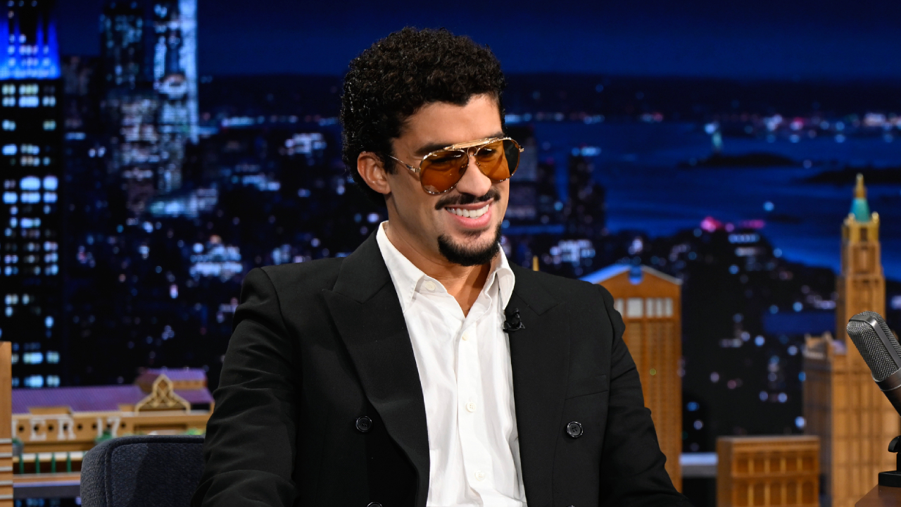 Bad Bunny with curly hair and sunglasses sits smiling on a talk show set, wearing a black jacket and white shirt, with a cityscape backdrop.