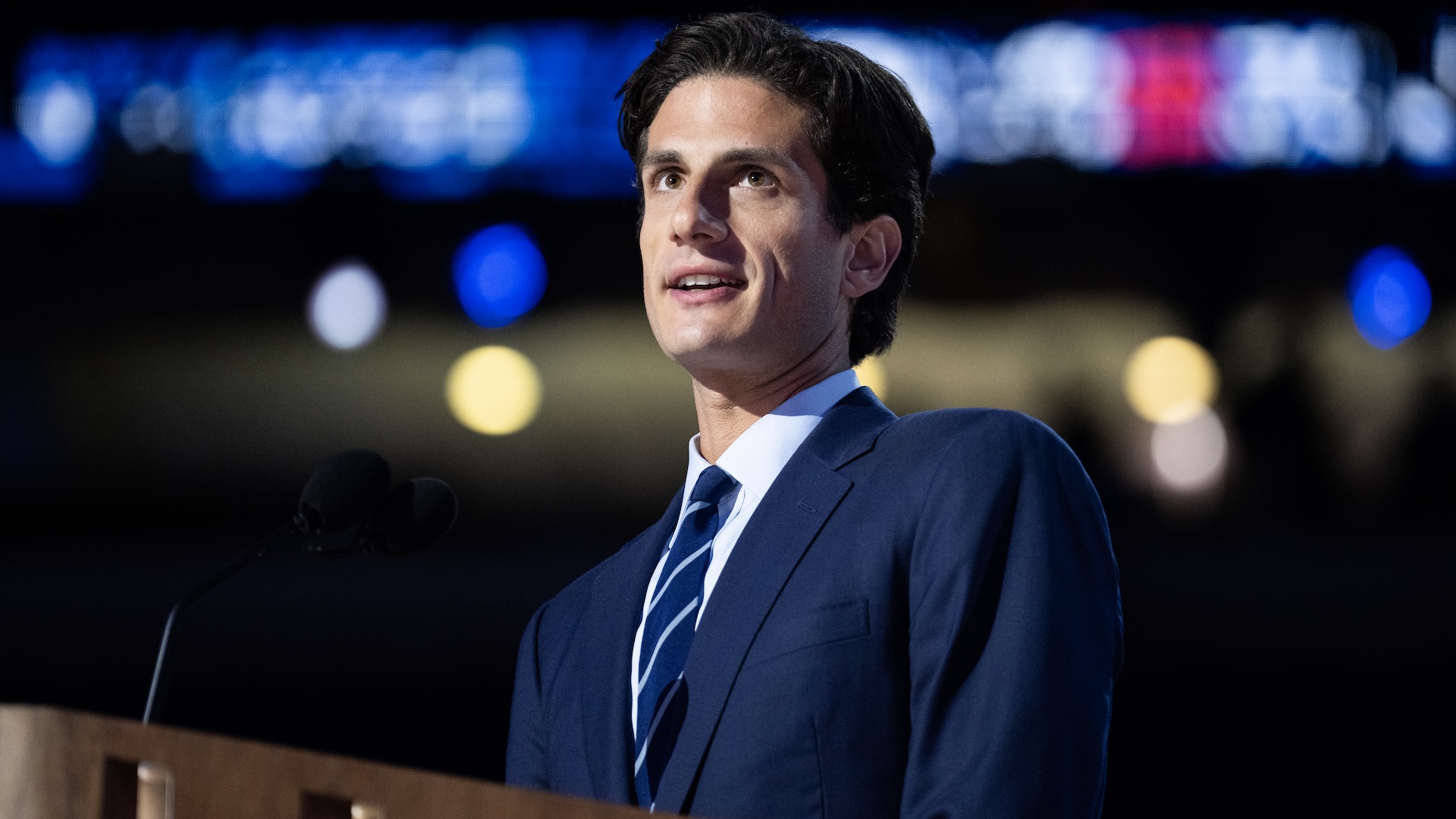 Jack Scholssberg in a suit speaks at a podium with blurred lights in the background.