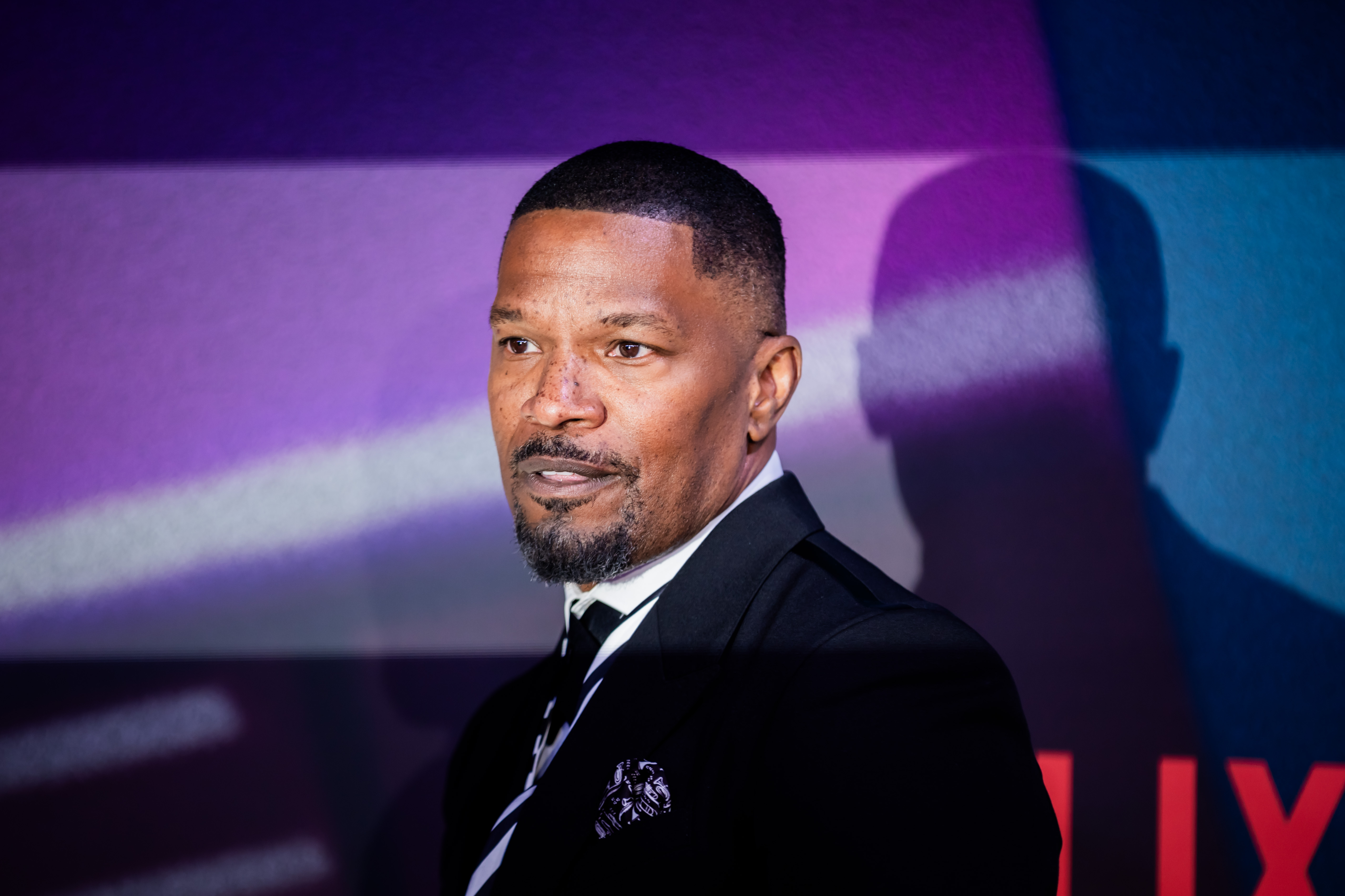 Jamie Foxx in a suit, standing against a colorful background, looking to the side with a slight smile.