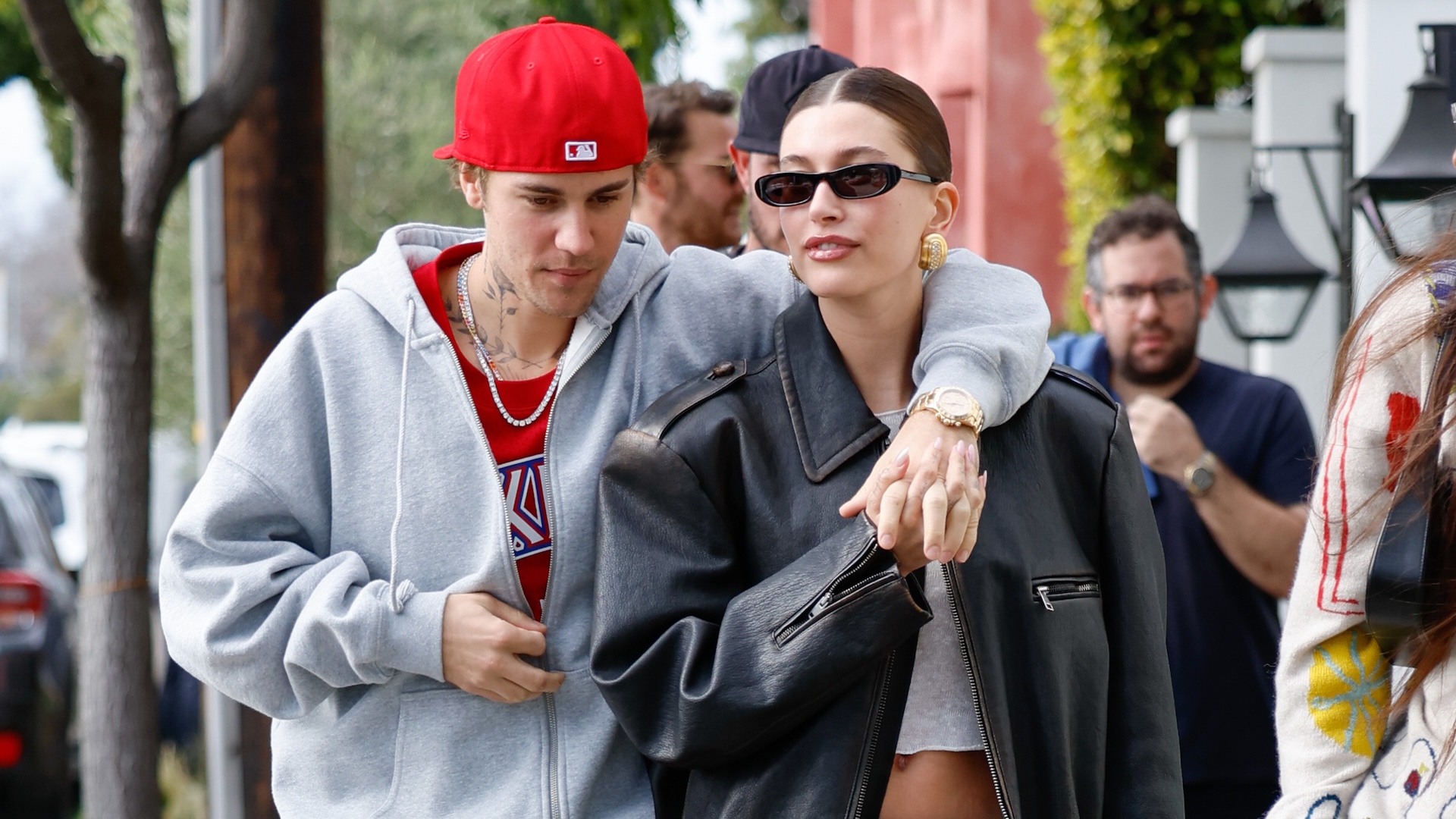 Justin Bieber in a red cap and grey hoodie, walking with Hailey Bieber in sunglasses and a black jacket. They are outdoors.