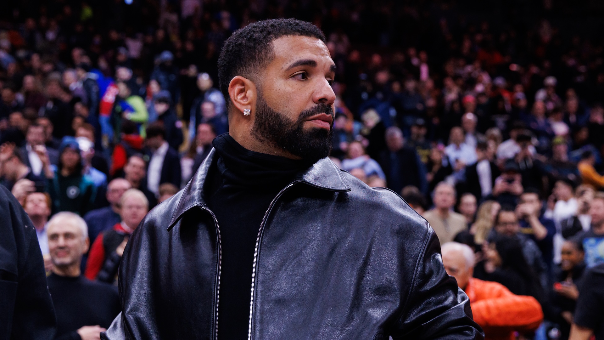 Drake, wearing a black leather jacket, stands in a crowded stadium, looking to the side.
