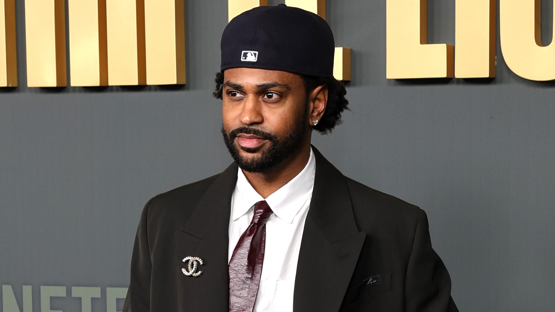 Big Sean wearing a suit with a Chanel brooch and a baseball cap, standing in front of a backdrop with gold lettering.
