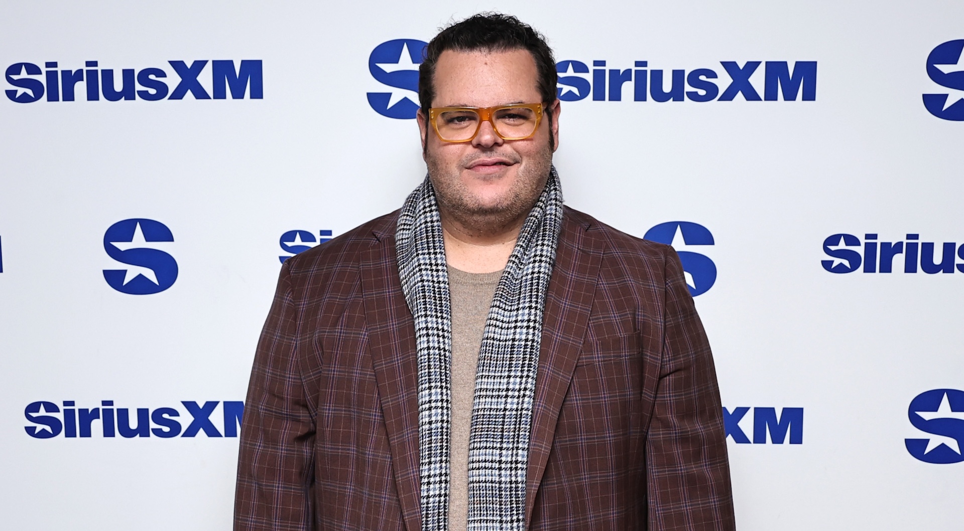 Josh Gad at a SiriusXM event, wearing a brown plaid jacket and scarf, with yellow-tinted glasses.