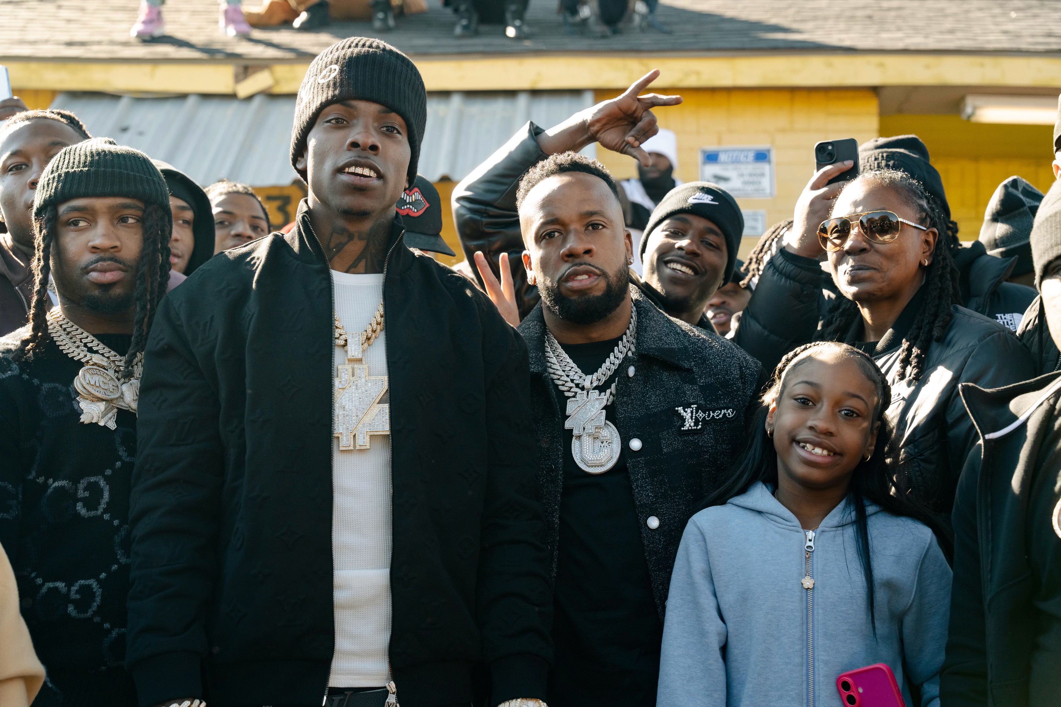 A group of people, including a young girl in a blue hoodie, stand together outdoors. Some wear black jackets and beanies, and there are smiles and gestures.