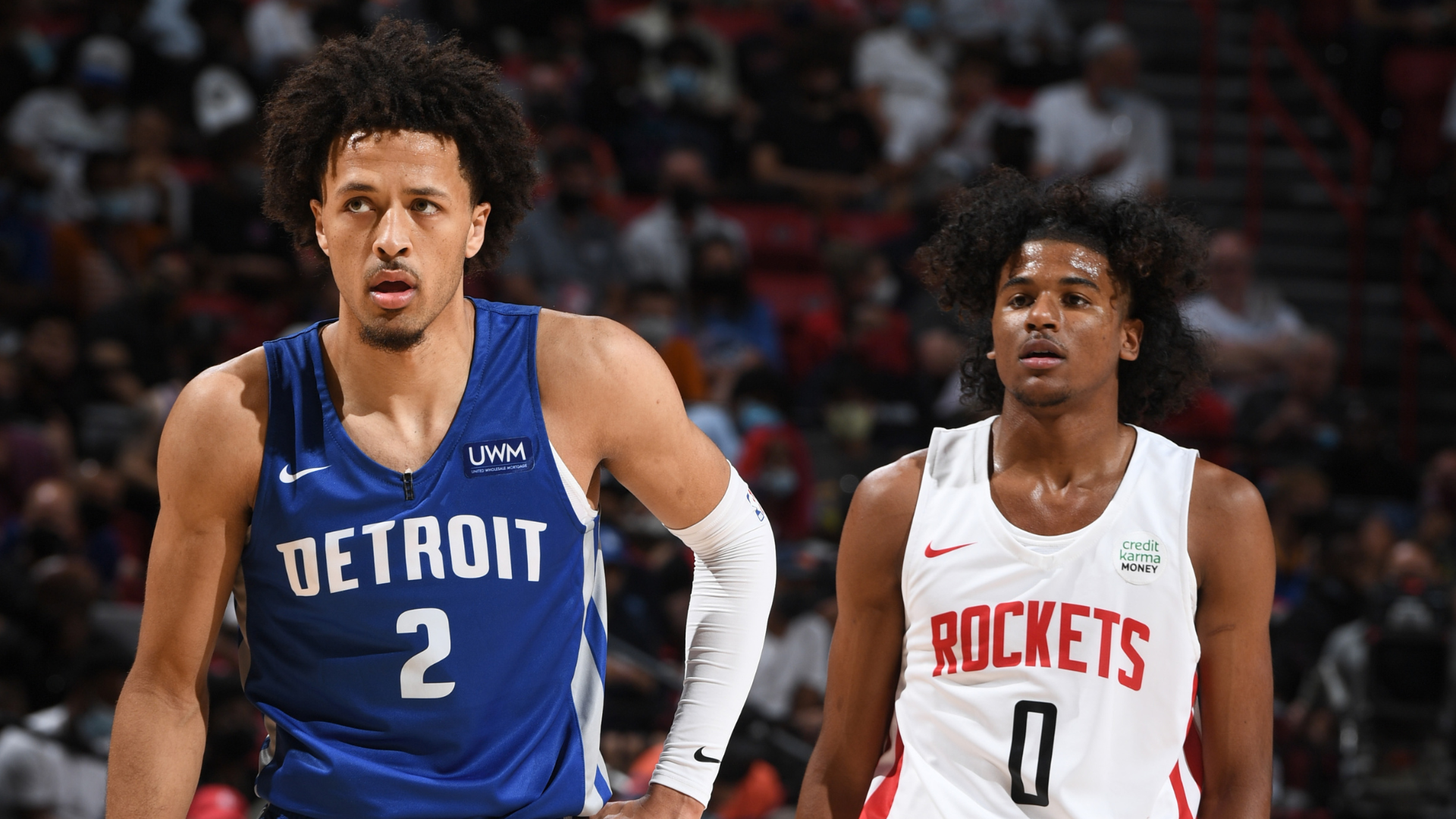 Two basketball players on the court: one in a blue "Detroit" jersey and the other in a white "Rockets" jersey, focused on the game.