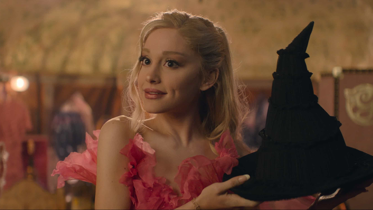 Ariana Grande as Glinda in 'Wicked' holding a witch hat, wearing a pink ruffled dress, with a whimsical expression in a warmly lit setting.