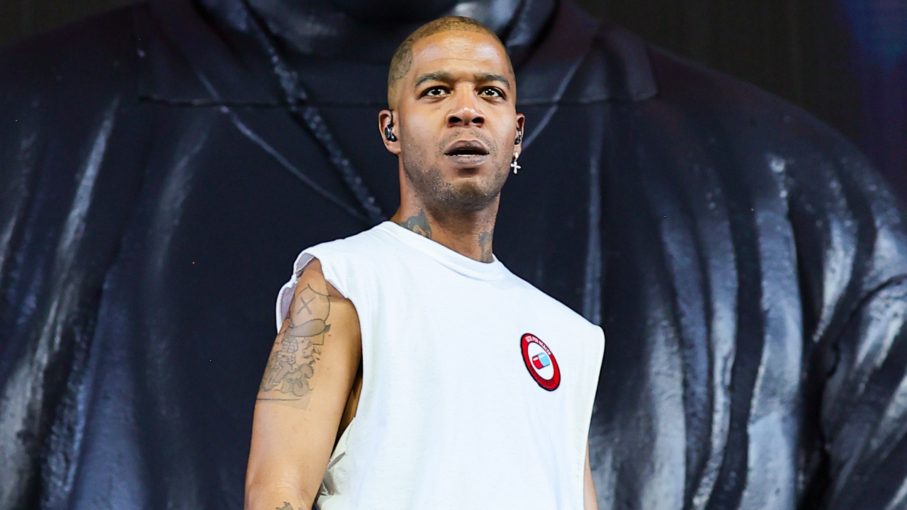 Kid Cudi performing on stage, wearing a sleeveless white shirt with tattoos visible on his arm, against a dark backdrop.