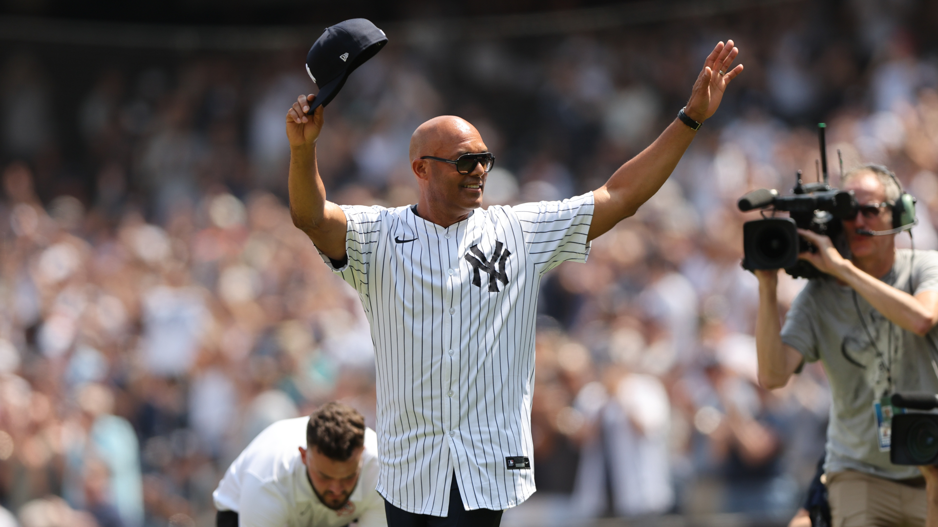 A man in a New York Yankees jersey waves his cap to a cheering crowd. A cameraman films the moment.