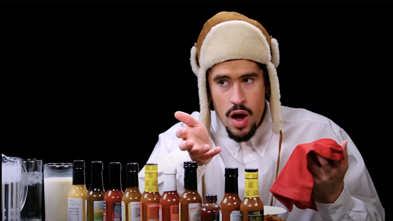 Bad Bunny in a winter hat sits at a table with various hot sauces, holding a red napkin.