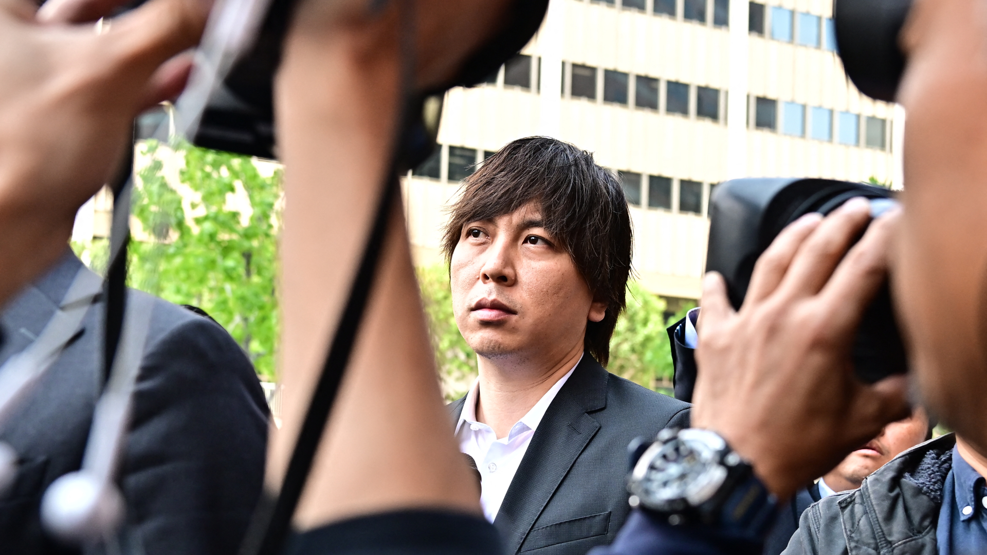 A man in a suit surrounded by photographers and cameras, with a city building in the background.